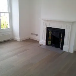 Jasnoszara podłoga z paneli ułożona w pustym pokoju z białymi ścianami, grzejnikiem pod oknem i ozdobnym kominkiem.