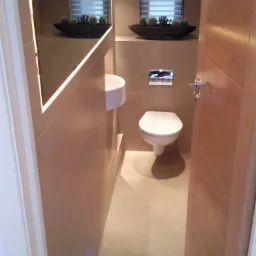 Kompaktowa toaleta w beżowych płytkach z podświetlanym lustrem, umywalką nablatową i dekoracyjnymi sukulentami, widok zza uchylonych drzwi.