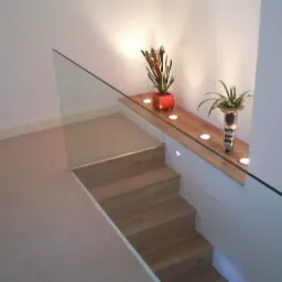 Jasne drewniane schody z szklaną balustradą, oświetlenie punktowe w półce z roślinami, minimalistyczny design.