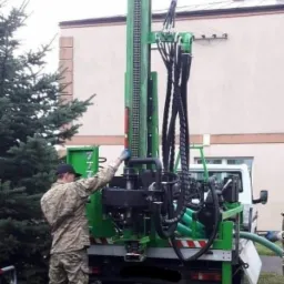 Pracownik w stroju moro obsługuje wiertnicę zamontowaną na samochodzie dostawczym, podczas wiercenia otworu w ziemi.