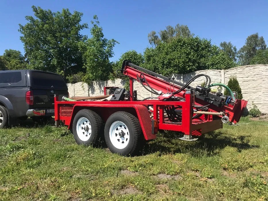 Czerwona wiertnica do studni głębinowych na przyczepie, podłączona do czarnego pickupa na tle zielonego trawnika i kamiennego muru.