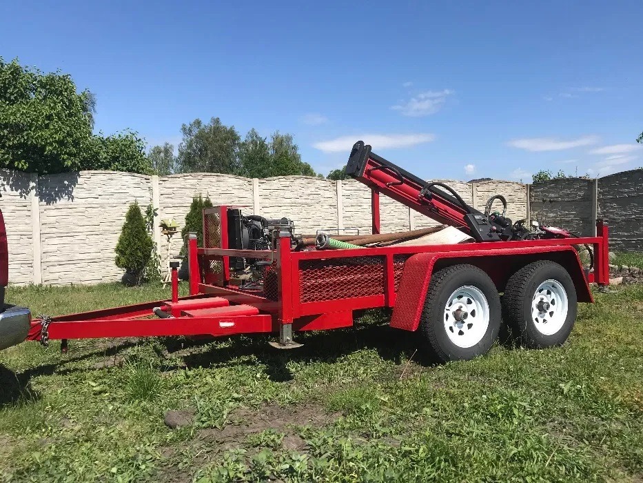 Czerwona przyczepa z wiertnicą do studni na trawiastym terenie, widoczny silnik, rury wiertnicze i fragment ogrodzenia z prefabrykatów betonowych w tle.