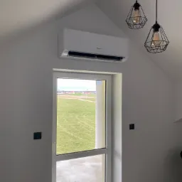 Biały klimatyzator Hisense zamontowany nad oknem w pokoju z widokiem na zielone pole, oświetlony nowoczesnymi lampami wiszącymi z metalowymi kloszami.