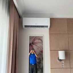 KLIMANEO Zbigniew Pigiel - Biały klimatyzator marki Gree zamontowany w pomieszczeniu z beżowymi zasłonami i tapicerowaną ścianą, odbicie instalatora w lustrze.