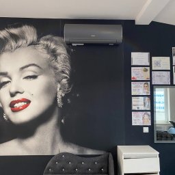 KLIMANEO Zbigniew Pigiel - Szary klimatyzator marki Gree zamontowany w salonie z fototapetą Marilyn Monroe, obok ściana z certyfikatami i szarym krzesłem z kryształkami.