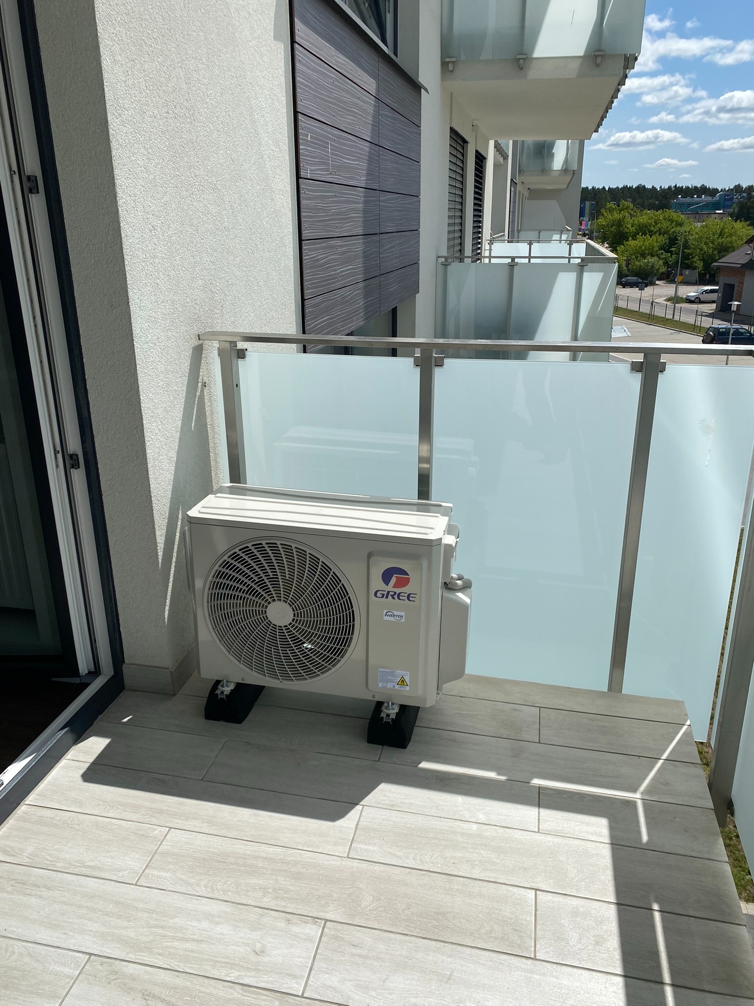 Kompaktowa jednostka zewnętrzna klimatyzacji marki Gree zamontowana na balkonie nowoczesnego apartamentu, widok z bliska, szklana balustrada, płytki imitujące drewno.