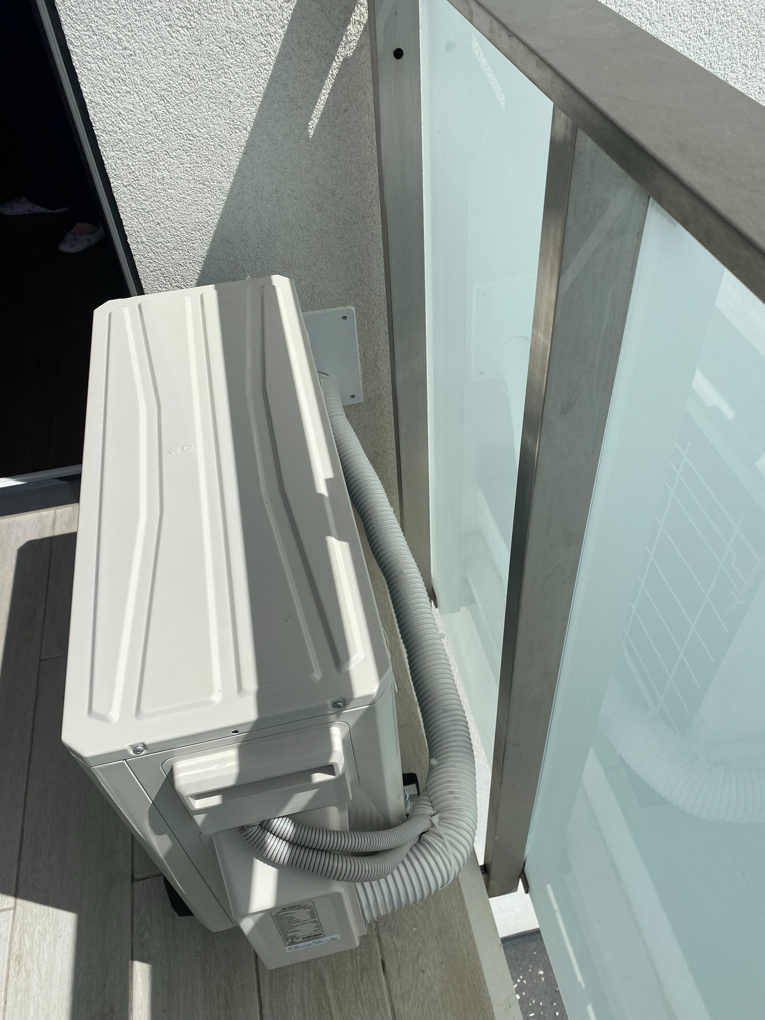 Zewnętrzna jednostka klimatyzacji zamontowana na balkonie z szklaną balustradą, widoczny przewód odprowadzający skropliny.