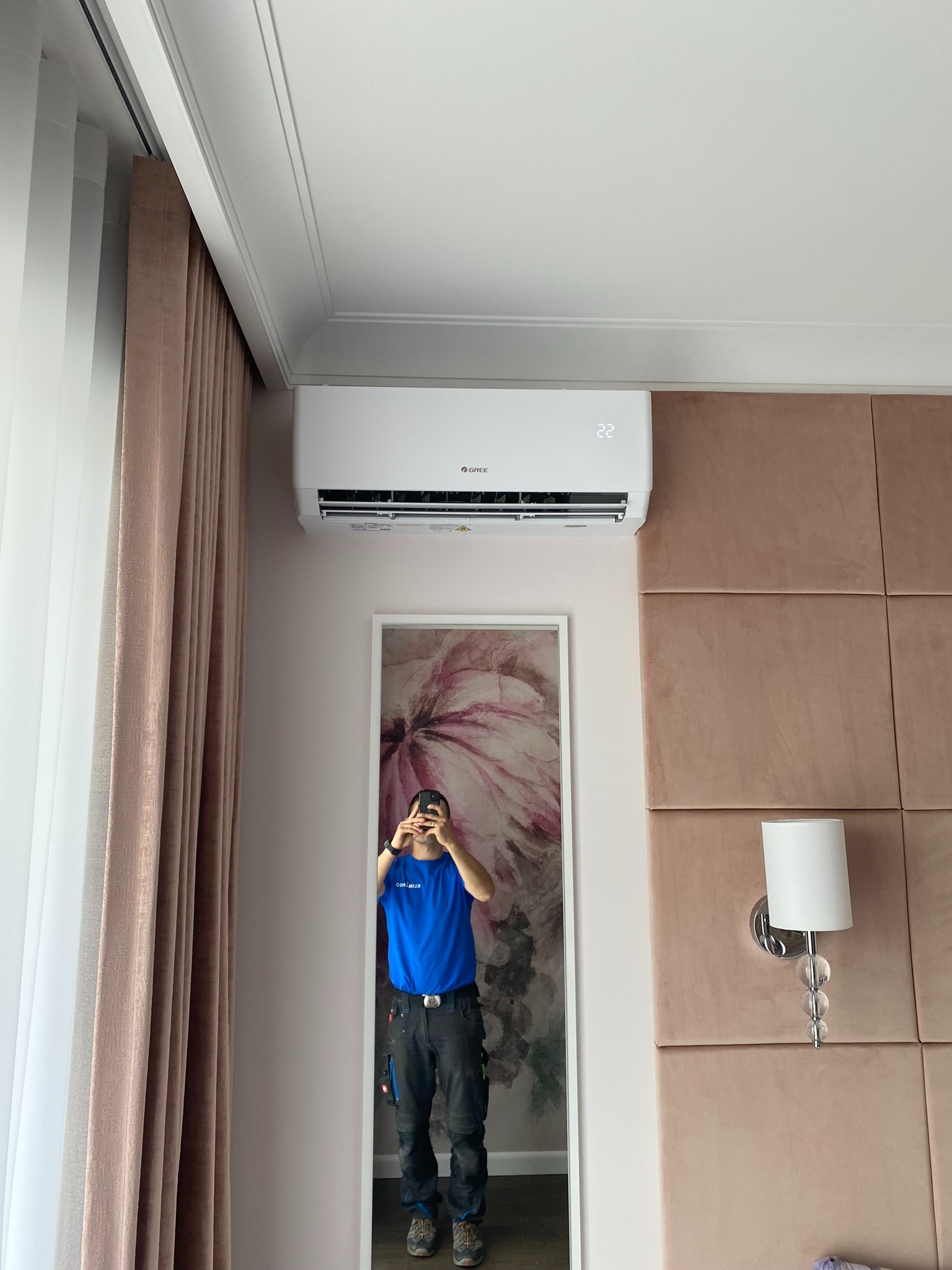 Biały klimatyzator marki Gree zamontowany w pomieszczeniu z beżowymi zasłonami i tapicerowaną ścianą, odbicie instalatora w lustrze.