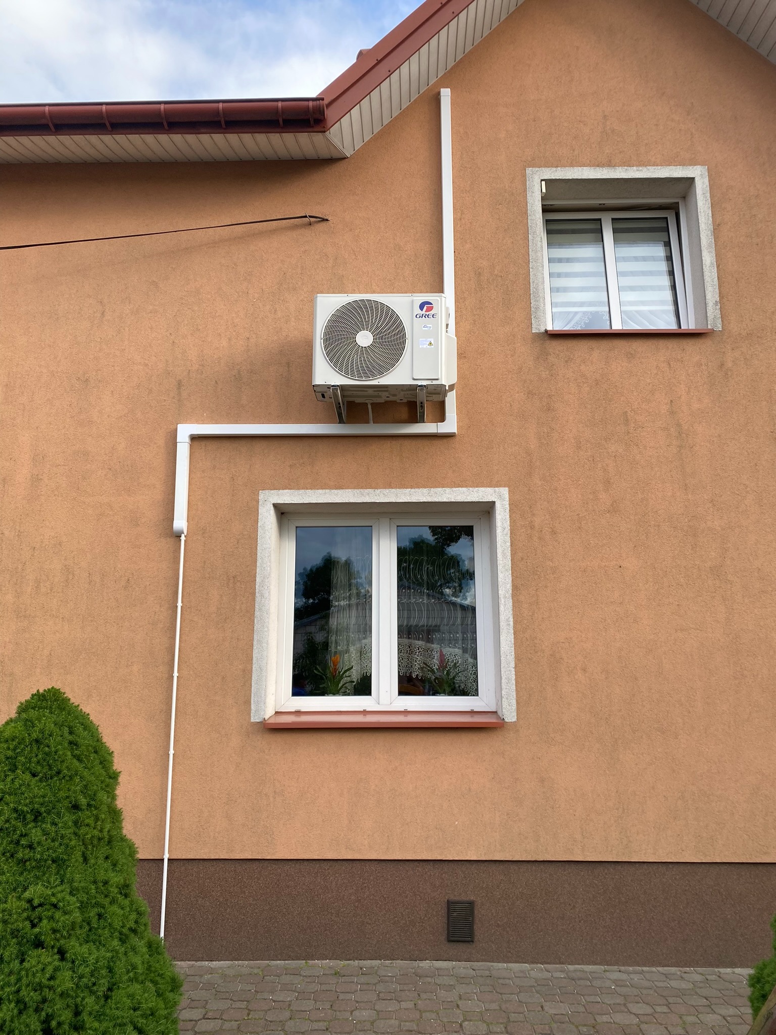 Zewnętrzna jednostka klimatyzacji marki Gree zamontowana na elewacji domu w kolorze brzoskwiniowym, widoczne białe rury odprowadzające skropliny, dwa okna z białymi ramami i zielony krzew iglasty u...