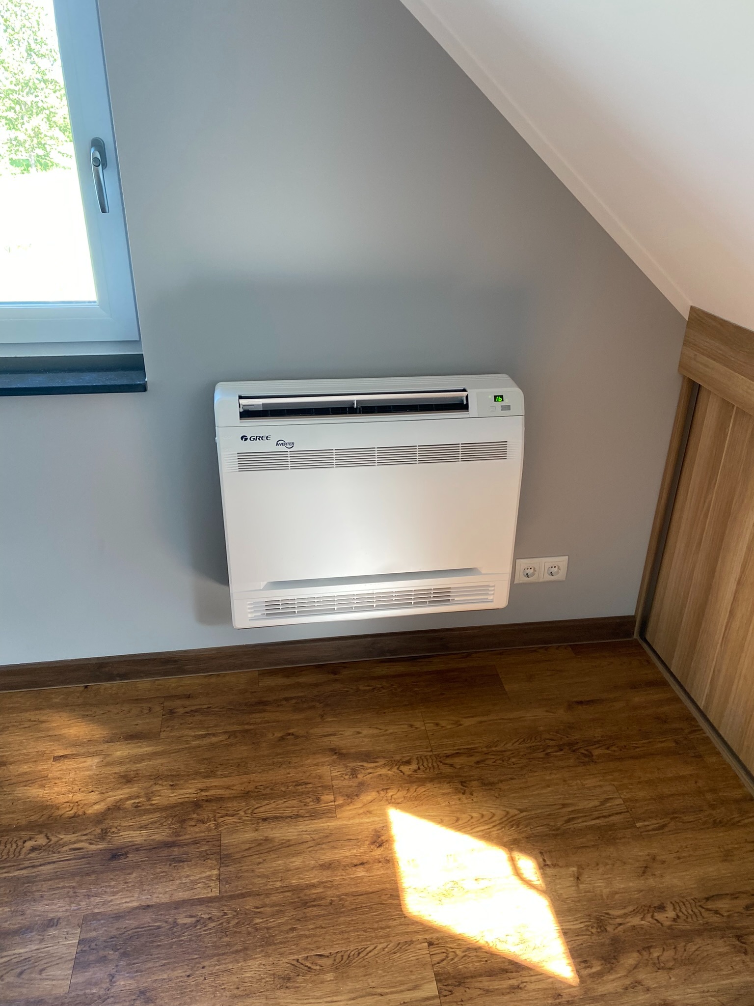 Biały klimatyzator podłogowy marki Gree zamontowany na szarej ścianie w pokoju na poddaszu, widoczne gniazdka elektryczne i fragment drewnianej szafy.