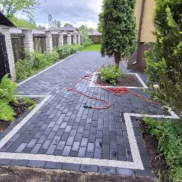 Ułożona, ciemnoszara kostka brukowa z jasnymi obwódkami, tworząca ścieżkę w ogrodzie z tujami i roślinami ozdobnymi, widoczny wąż ogrodowy.