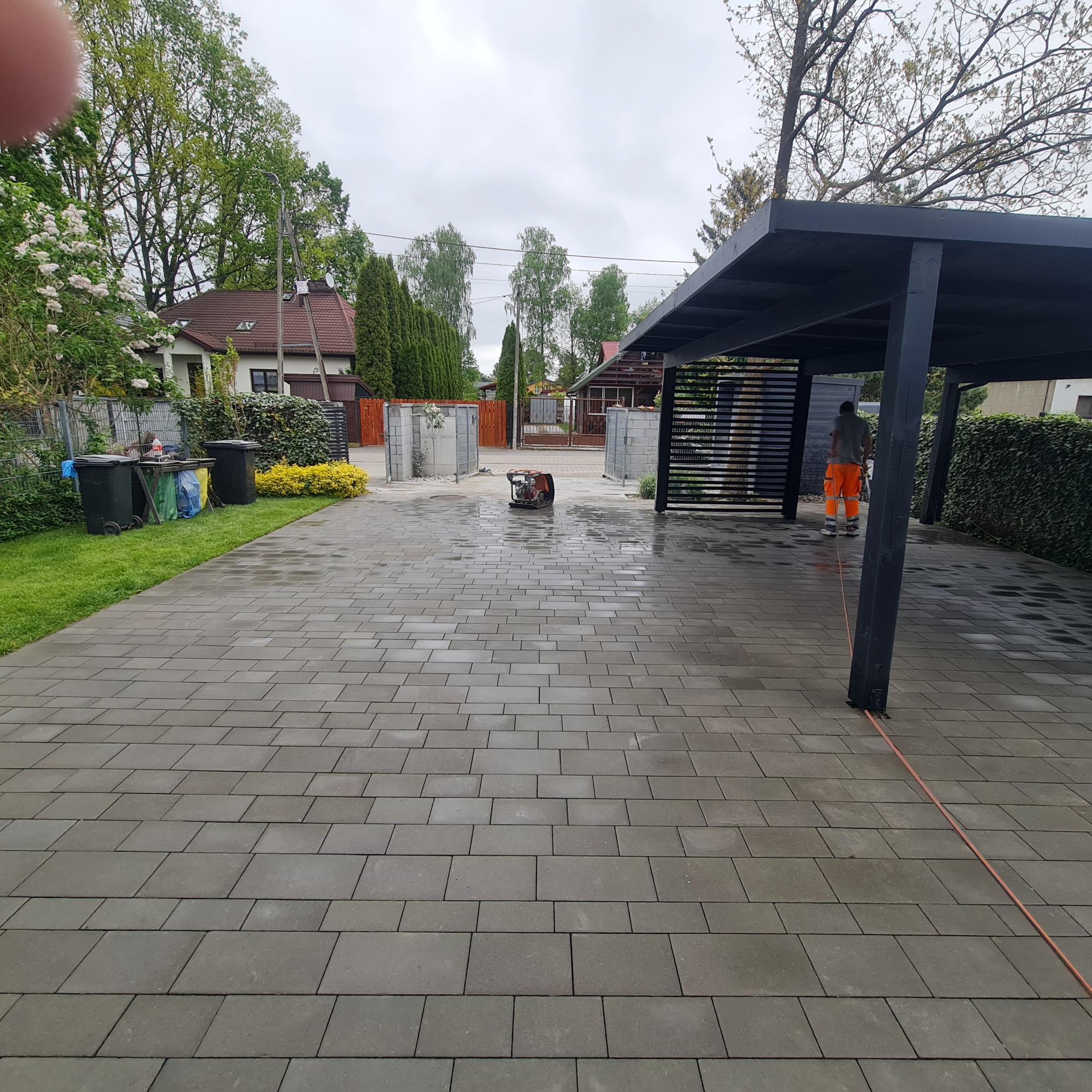 Świeżo ułożona, mokra kostka brukowa w odcieniach szarości na podjeździe, z widocznym pracownikiem w pomarańczowych spodniach pod zadaszeniem i zagęszczarką w tle.