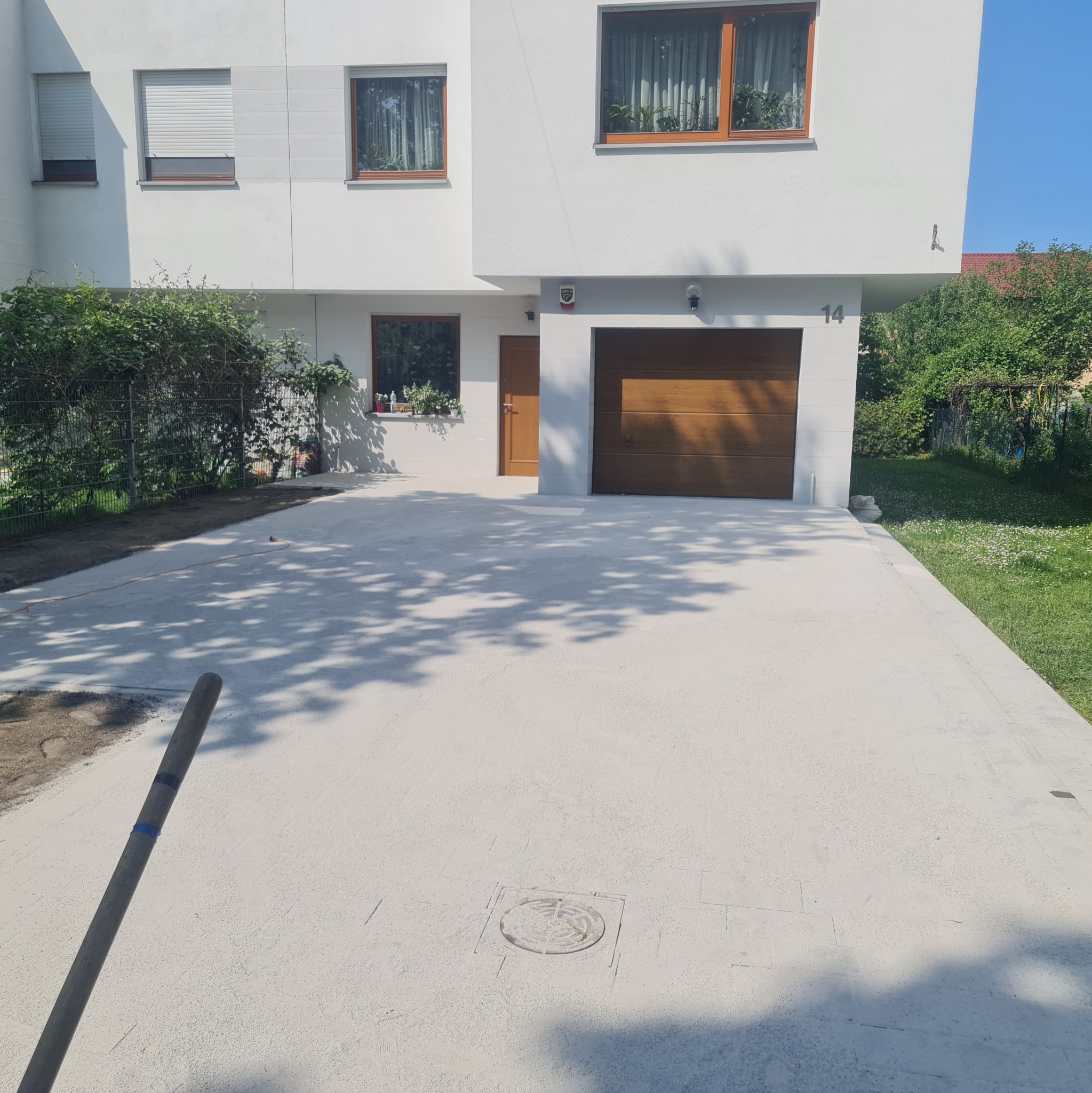 Nowa, jasna nawierzchnia z kostki brukowej przed nowoczesnym domem z drewnianą bramą garażową i numerem 14, widoczne cienie drzew i fragment narzędzia brukarskiego.