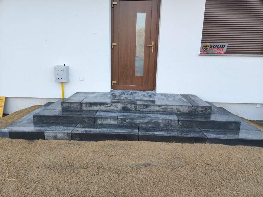 Schody wejściowe do domu wyłożone ciemnoszarą kostką brukową o fakturze drewna, otoczone piaskiem, z brązowymi drzwiami i roletami w tle.