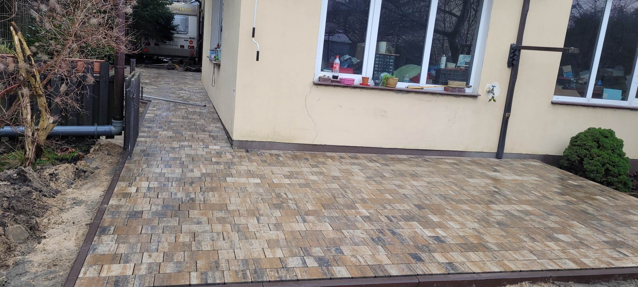 Nowo ułożona, wielobarwna kostka brukowa prowadząca do budynku z beżową elewacją, widoczne okno z roślinami na parapecie i fragment ogrodzenia z rurą odpływową