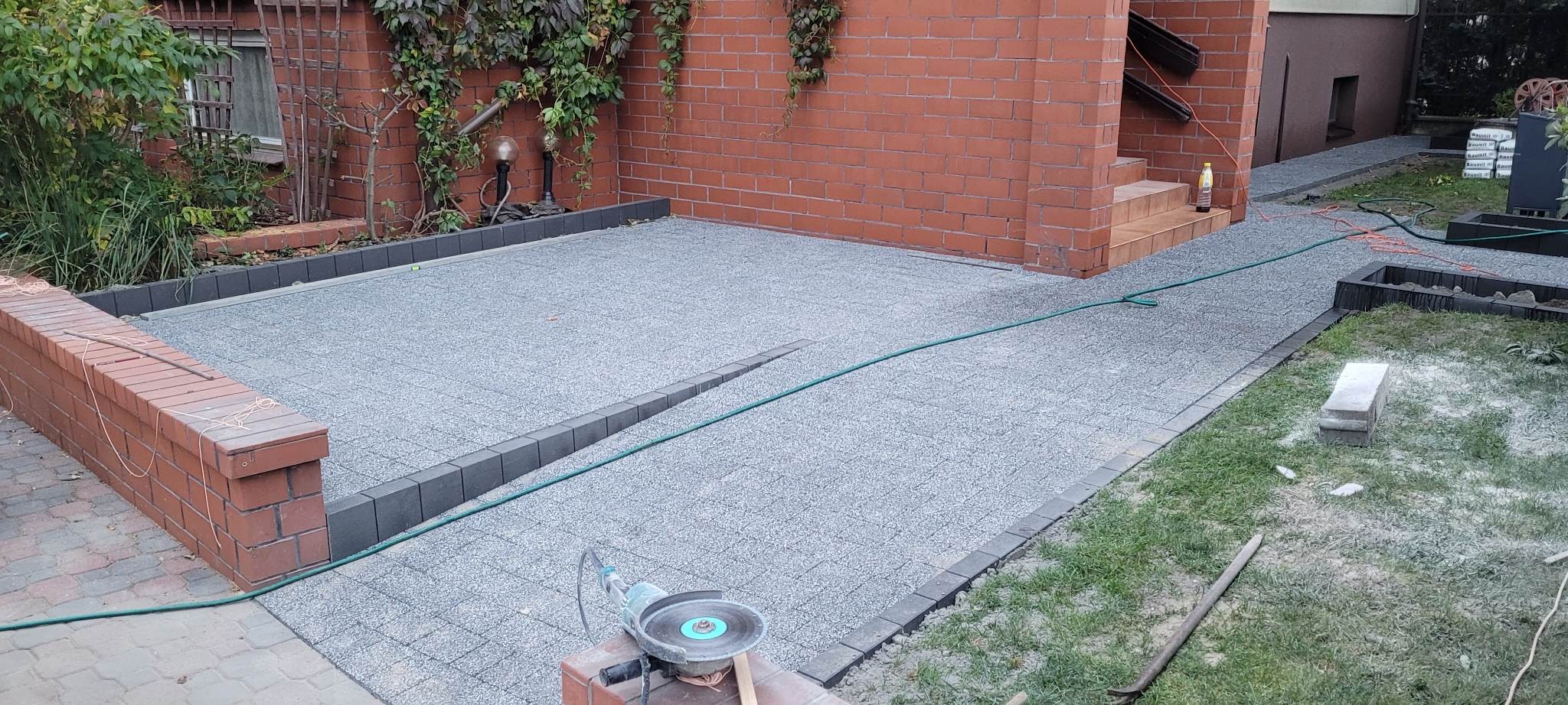 Niedokończona nawierzchnia z szarej kostki brukowej z czarnym obrzeżem, widoczna szlifierka kątowa na ceglanym murku, zielony wąż ogrodowy i ceglany budynek w tle.