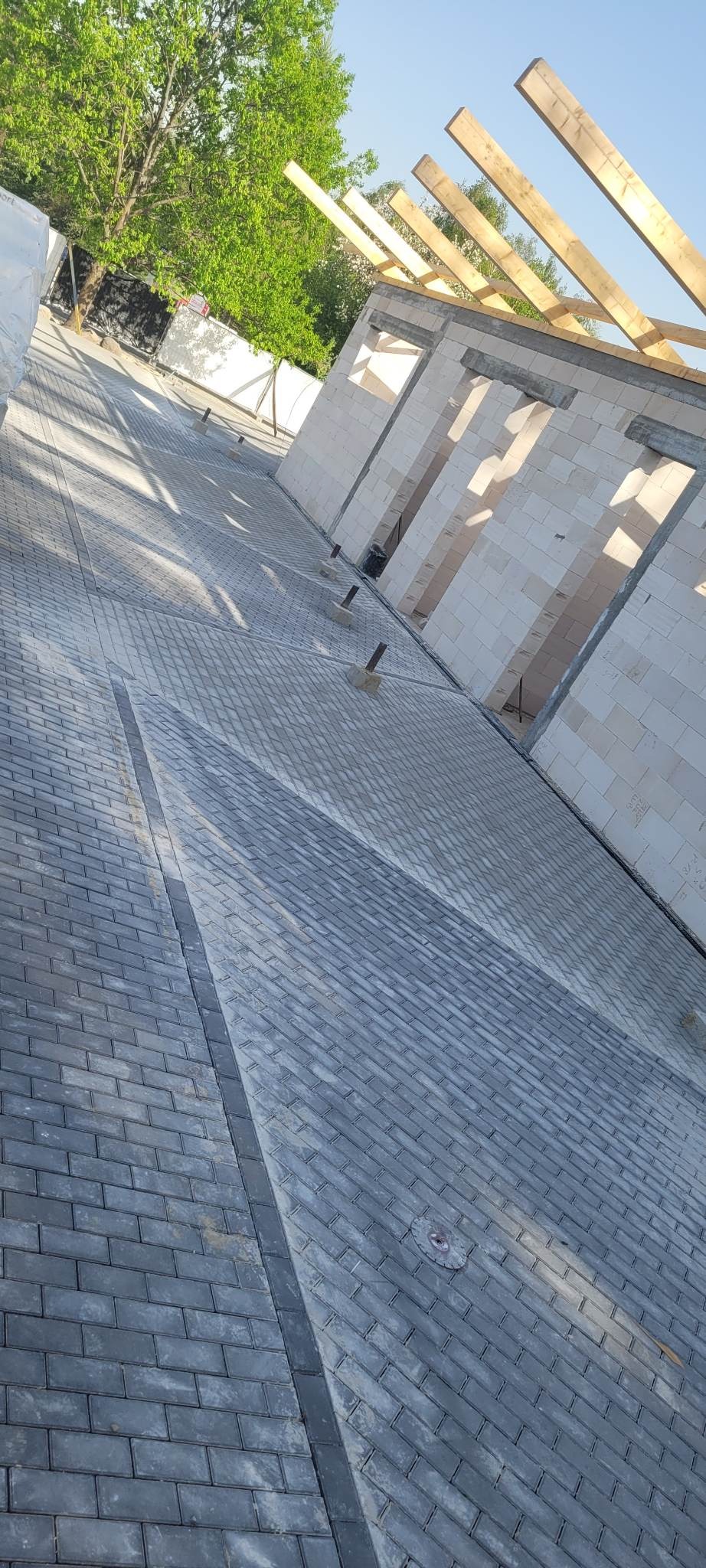 Ułożona szara kostka brukowa na podjeździe, z widocznym fragmentem budynku w trakcie budowy z drewnianą konstrukcją dachu, w słoneczny dzień.