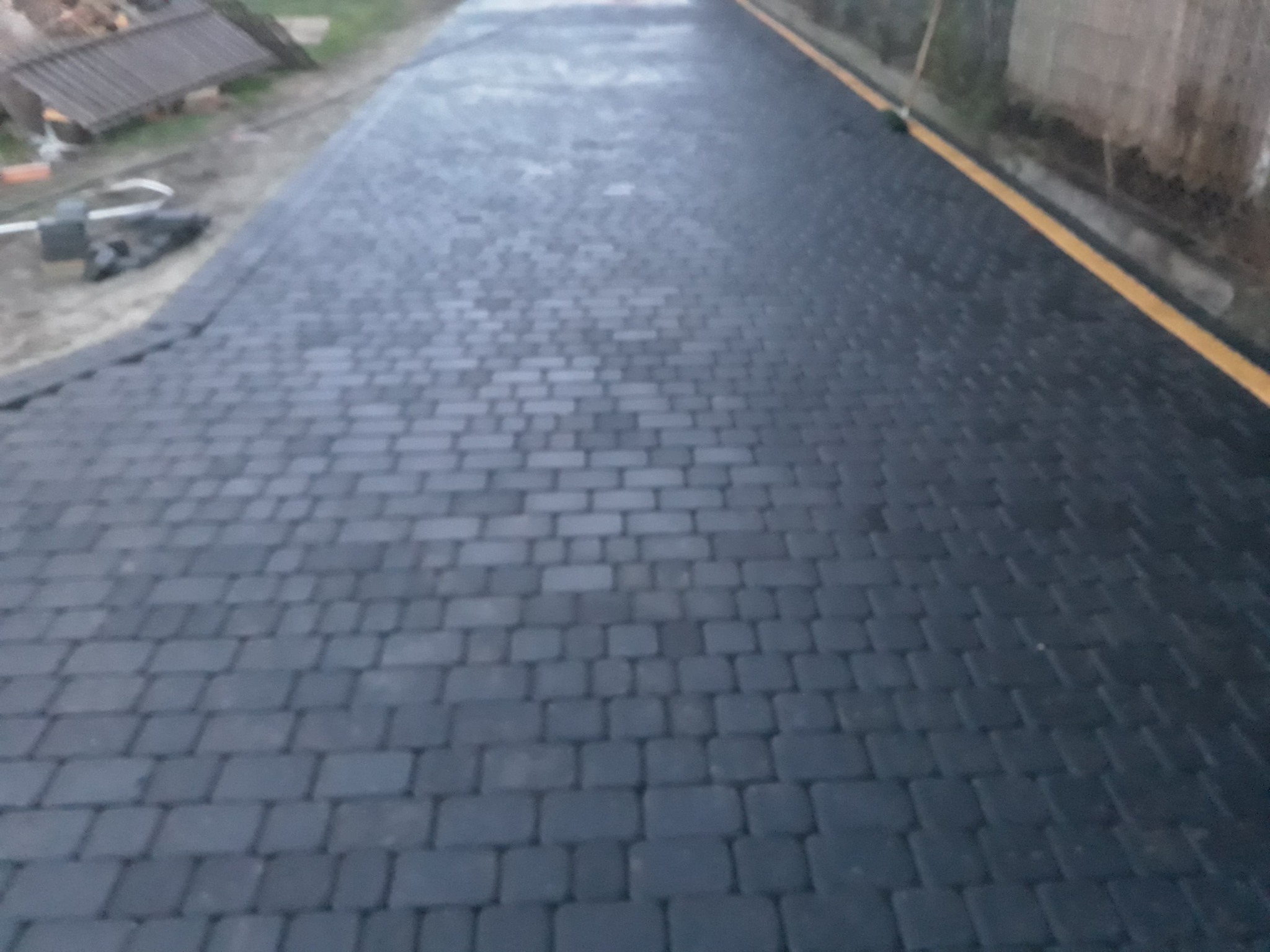 Ułożona, ciemnoszara kostka brukowa na podjeździe, widoczna żółta linia oddzielająca od trawnika, w tle częściowo widoczne materiały budowlane.