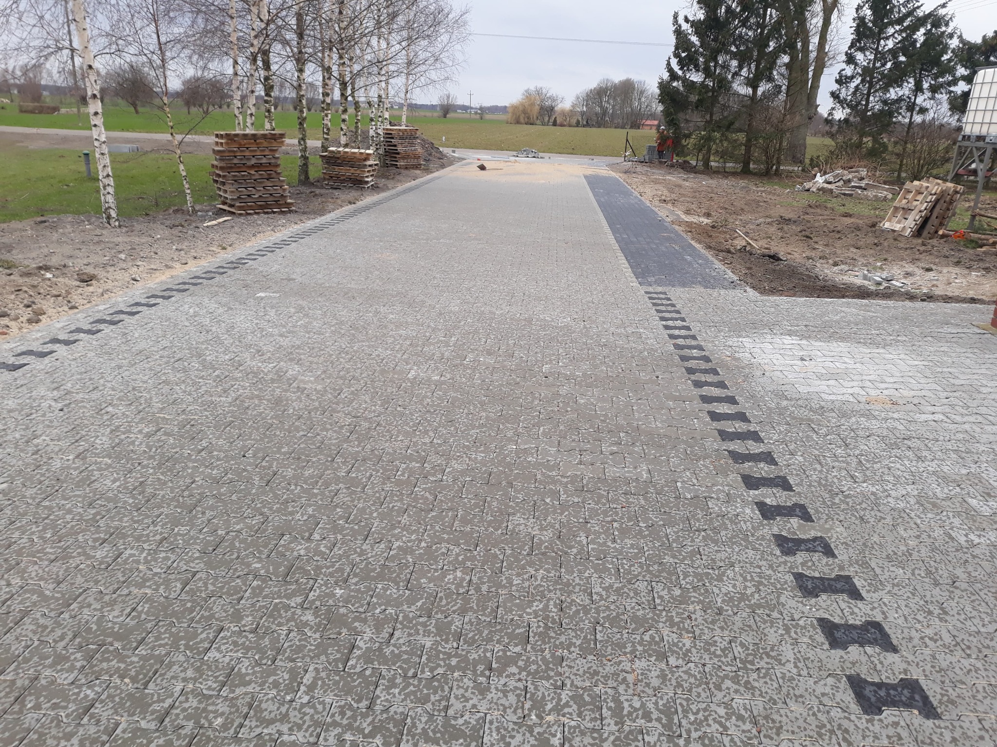 Ułożona szara kostka brukowa z czarnymi akcentami, palety kostki przy drzewach, teren w trakcie prac ziemnych w tle.
