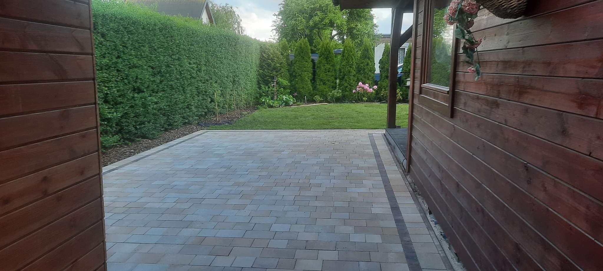 Ułożona kostka brukowa w odcieniach szarości i beżu, otoczona zielonym trawnikiem i wysokim żywopłotem, widok zza drewnianej ściany budynku.