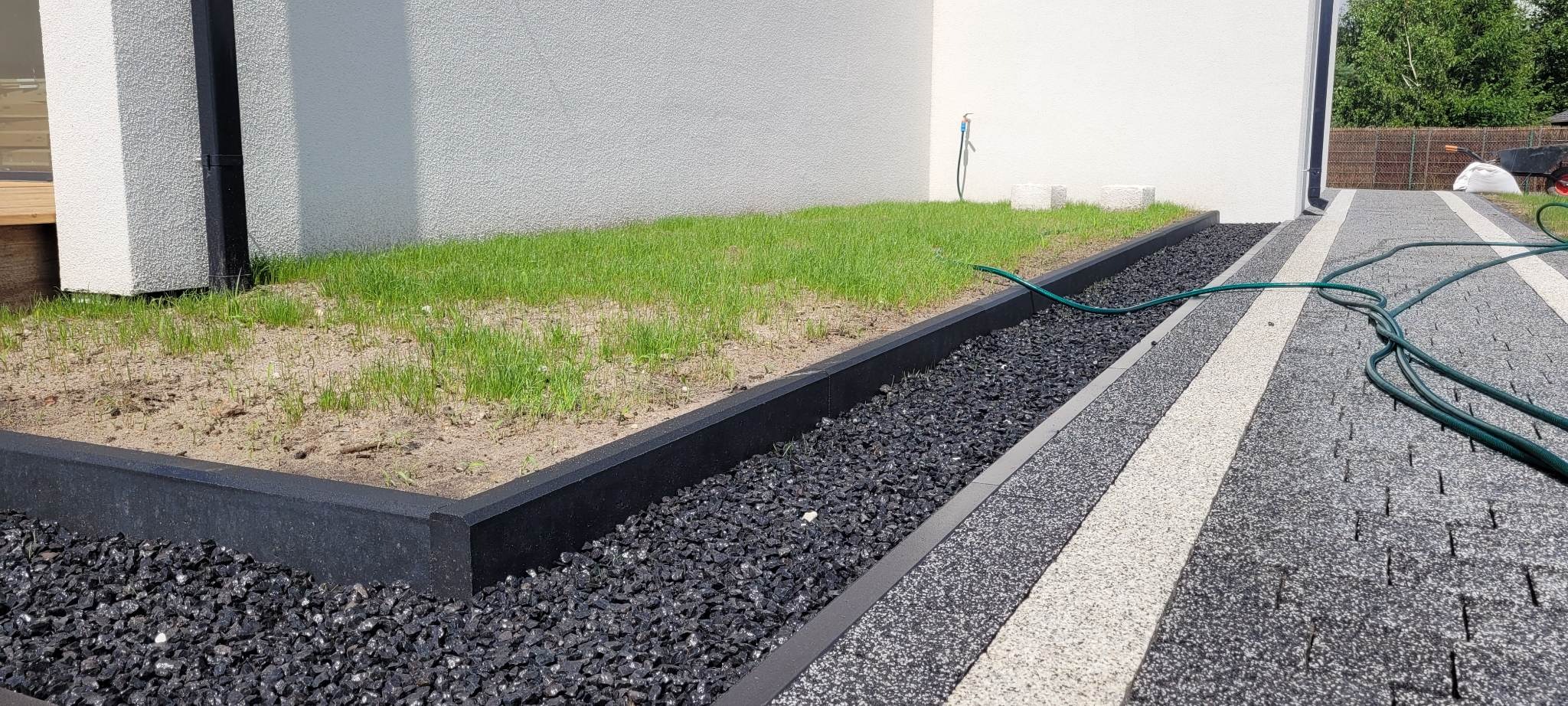 Ułożona kostka brukowa z czarną obwódką z kamieni ozdobnych, trawnik z młodą trawą i zielony wąż ogrodowy na posesji.