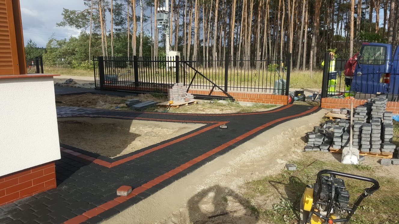 Częściowo ułożony chodnik z kostki brukowej w kolorze czarnym z pomarańczowymi paskami, prowadzący od domu do czarnej metalowej bramy wjazdowej. W tle widoczny las i niebieski samochód dostawczy,...