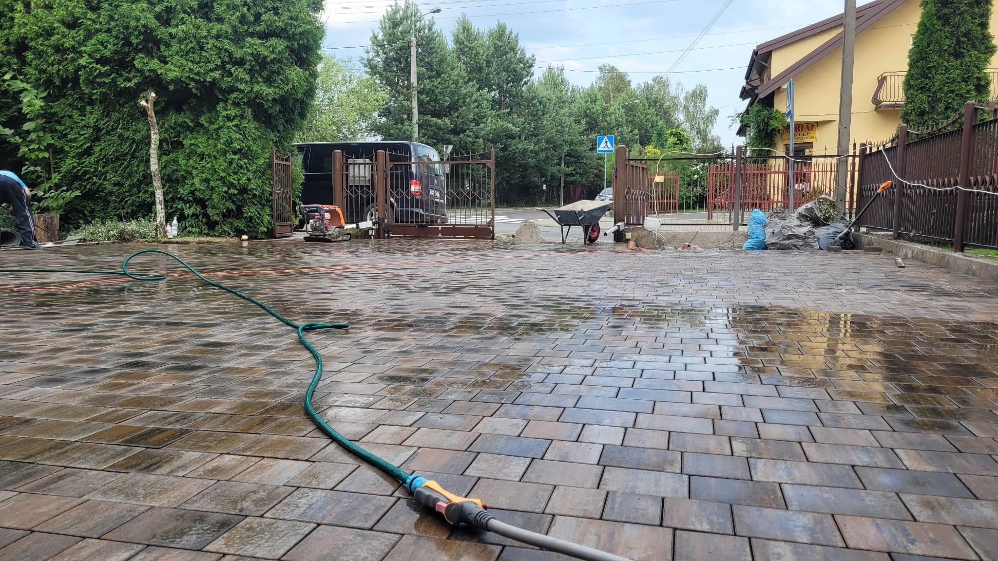 Mokry, brązowo-szary podjazd z kostki brukowej po czyszczeniu, z widocznym wężem ogrodowym, taczką i workami na śmieci w tle, częściowo widoczny budynek z żółtą elewacją.