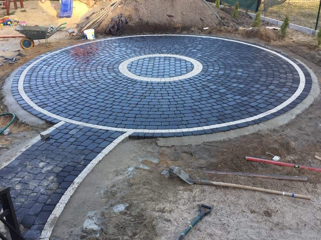 Okrągły plac z ciemnej kostki brukowej z białym obramowaniem, w trakcie układania, widoczne narzędzia i piasek.