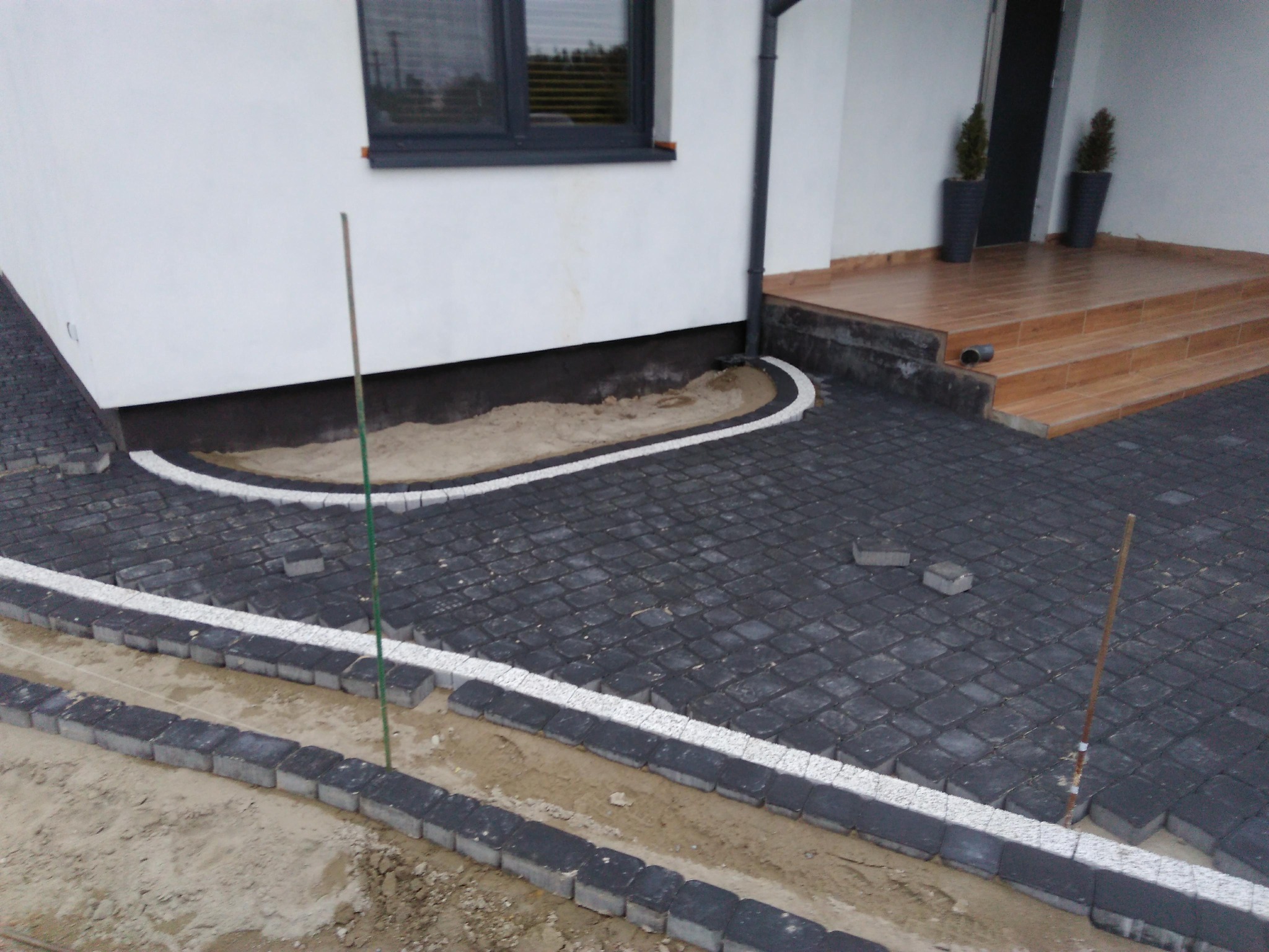 Częściowo ułożona, czarno-biała kostka brukowa przed domem z drewnianymi schodami i donicami z roślinami; widoczne etapy prac brukarskich, słupki wyznaczające poziom.