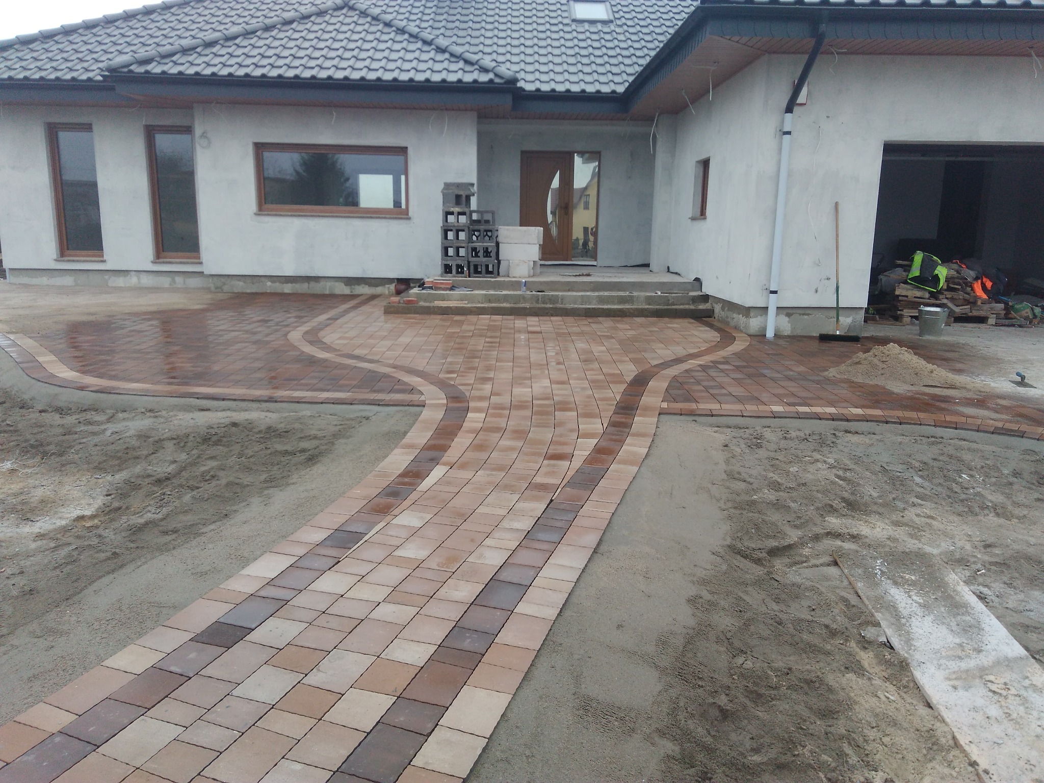 Ułożona, dwukolorowa kostka brukowa prowadząca do wejścia do domu z szarą elewacją i grafitowym dachem, widoczne przygotowane podłoże z piasku.