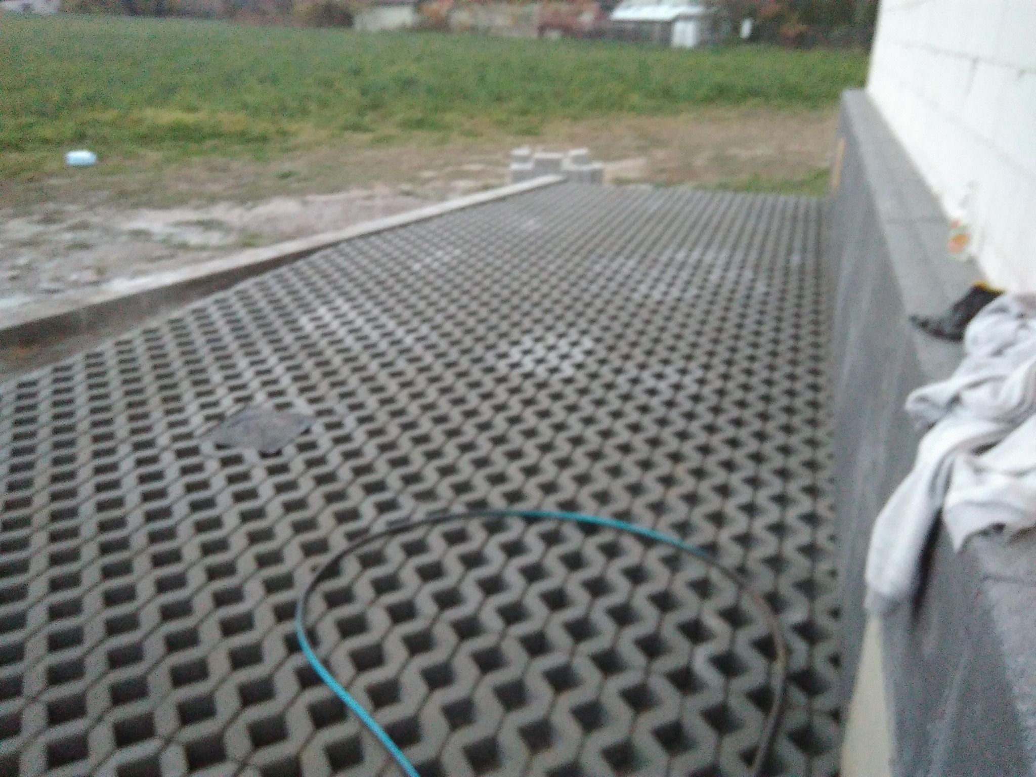 Szara ażurowa nawierzchnia z betonu na podwórku, widoczny fragment niebieskiego węża, ściana budynku i trawnik w tle.