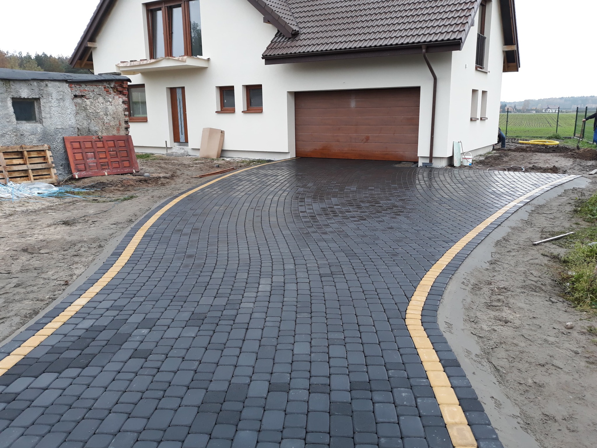 Czarno-żółta kostka brukowa ułożona w łuk przed domem z brązową bramą garażową, widoczne prace ziemne po bokach i elementy starego budynku w tle.
