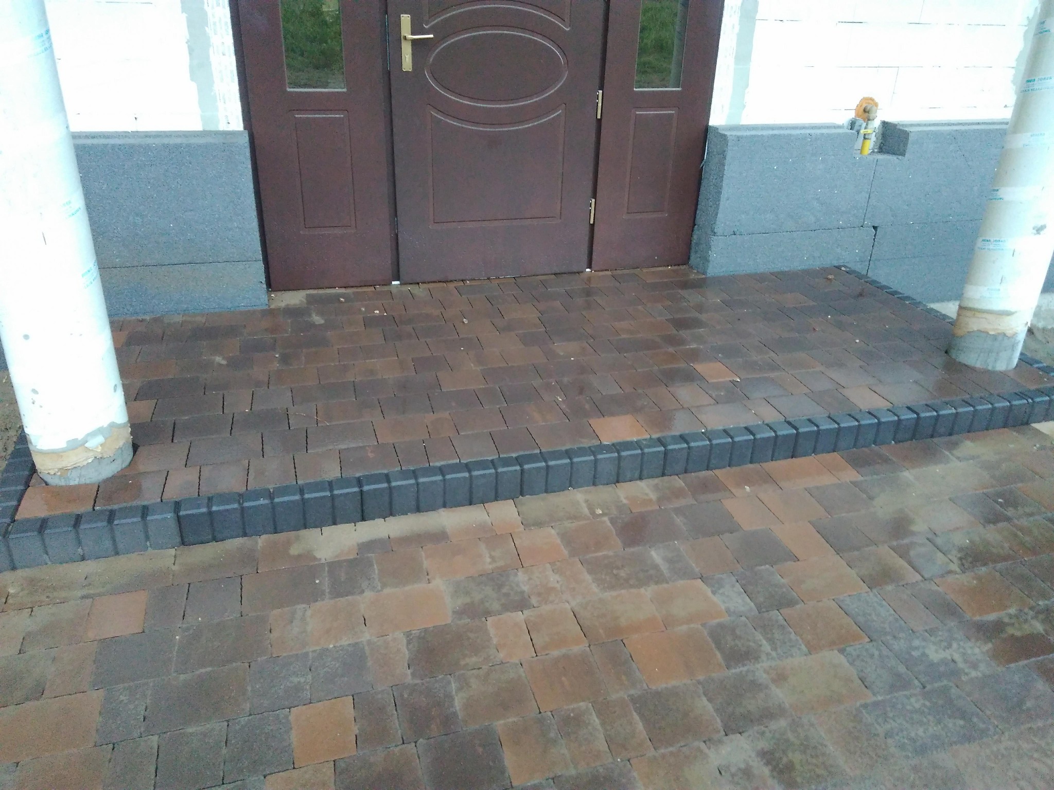Ułożona kostka brukowa w odcieniach brązu i szarości przed wejściem do budynku z brązowymi drzwiami, obrzeża z ciemnoszarej kostki, widoczne fragmenty kolumn i ocieplenia budynku.
