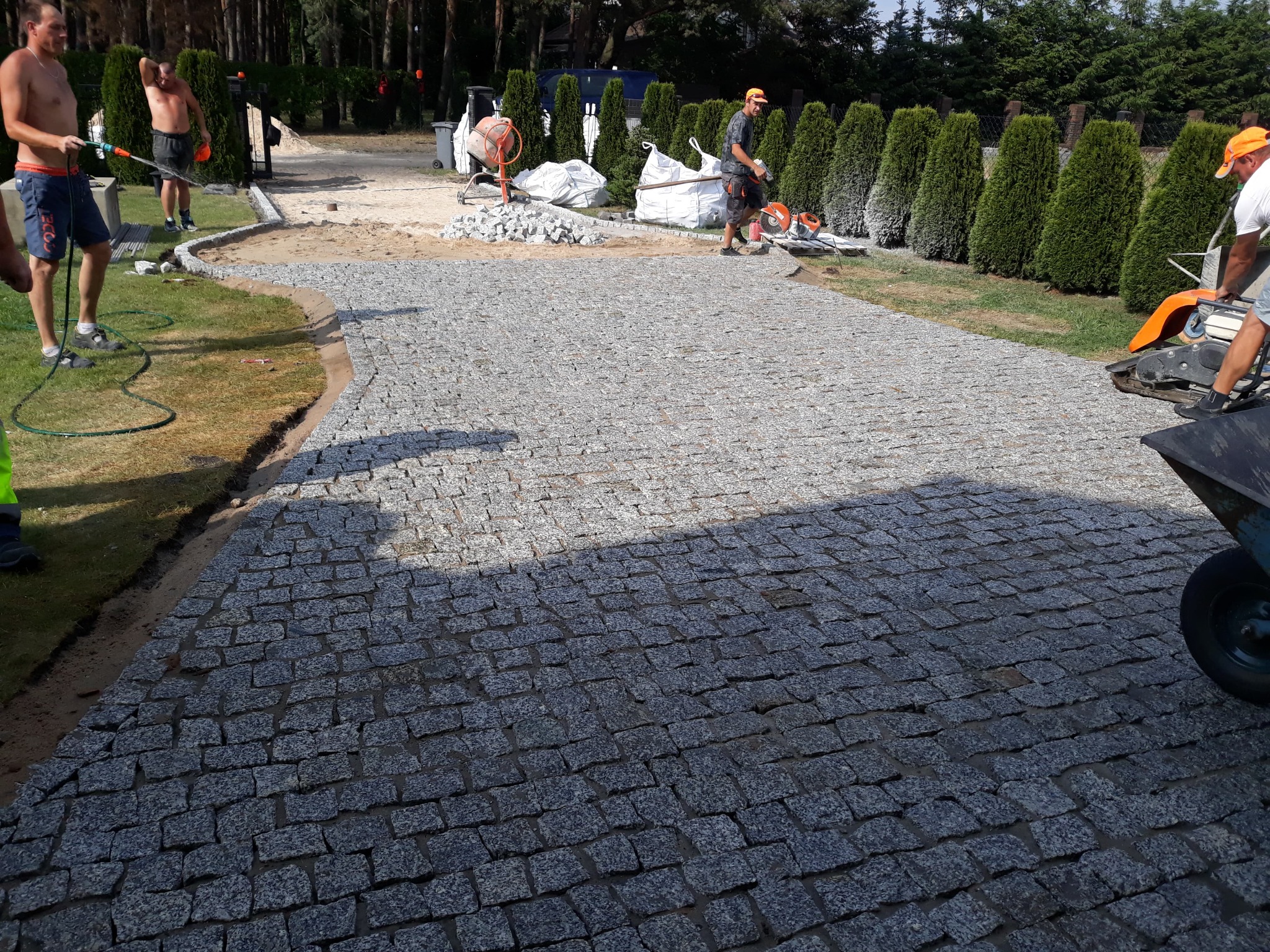 Trwa układanie szarej, granitowej kostki na podjeździe; widoczni robotnicy, betoniarka i narzędzia.