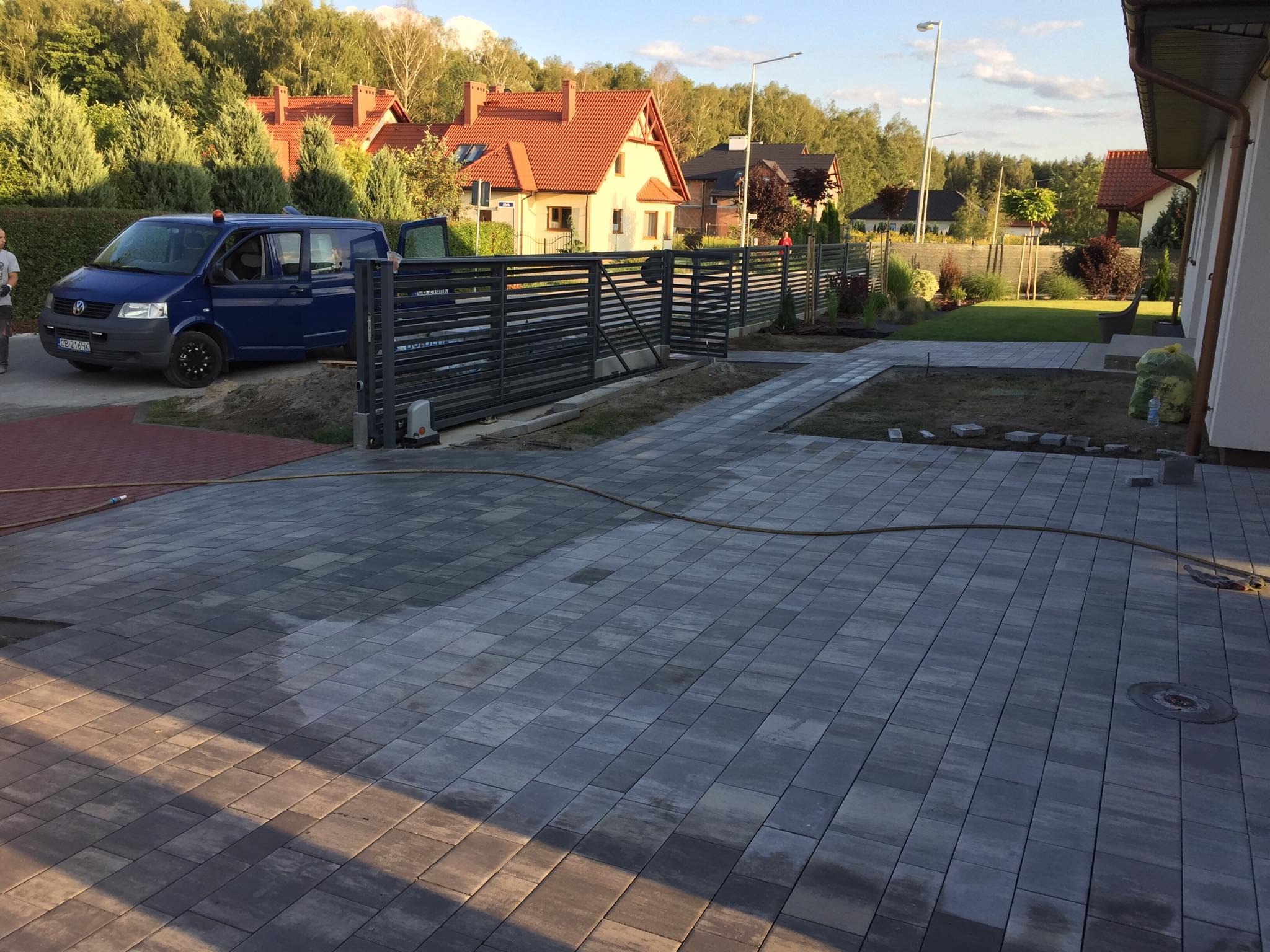 Ułożona szara kostka brukowa na podjeździe domu jednorodzinnego, z widocznym fragmentem metalowej bramy przesuwnej, niebieskim busem i elementami trawnika w trakcie aranżacji.