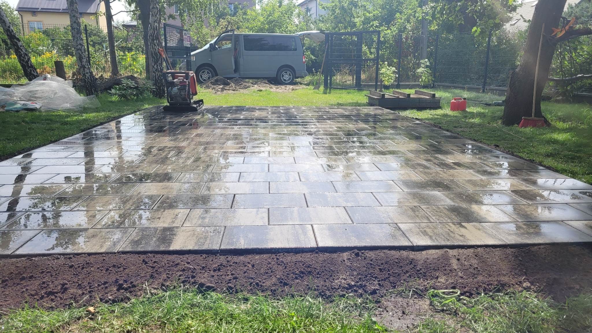 Świeżo ułożona, wilgotna kostka brukowa na przygotowanym podłożu z ubijarką w tle, widoczny samochód dostawczy i narzędzia ogrodowe.