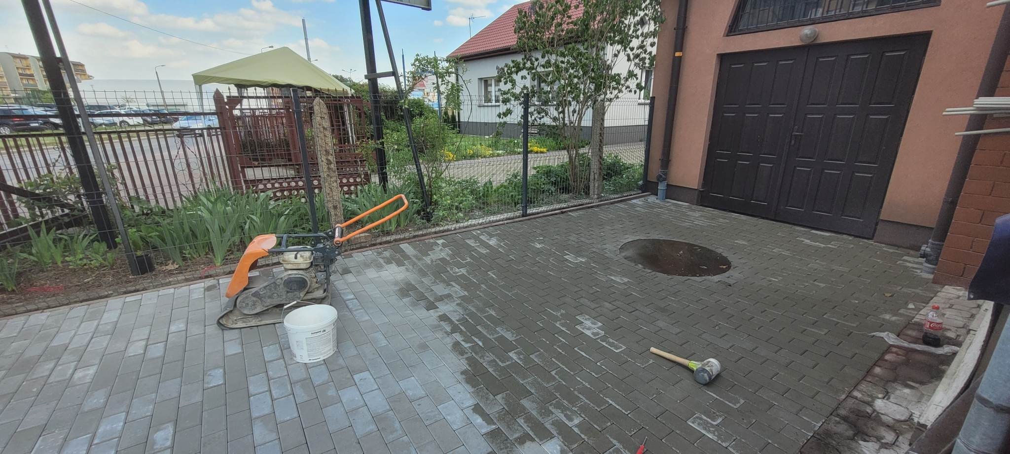 Ułożona szara kostka brukowa na podjeździe z widoczną ubijarką wibracyjną, gumowym młotkiem, białym wiadrem i kałużą. W tle metalowe ogrodzenie i budynek z bramą garażową.