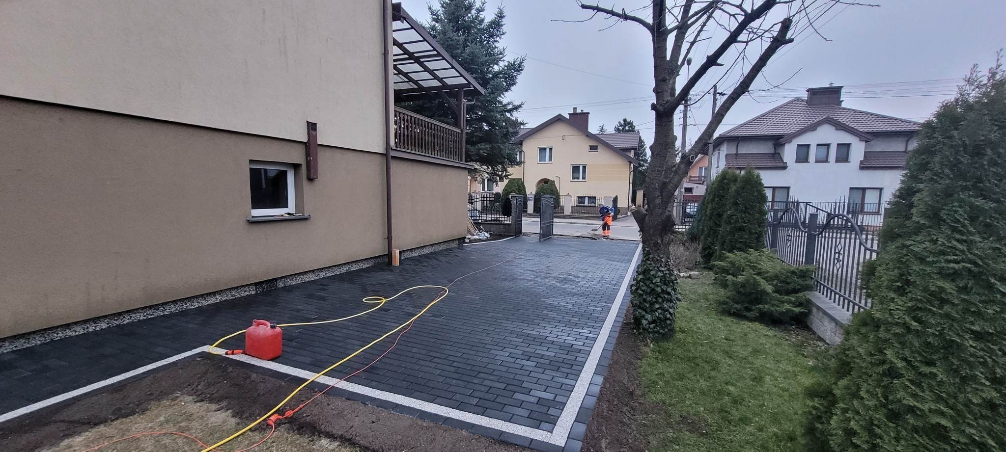 Ułożona, ciemnoszara kostka brukowa z jasnymi obrzeżami na podjeździe przed domem. W tle widoczny pracownik w pomarańczowym ubraniu podczas zamiatania. Czerwony kanister i żółty kabel leżą na kostce.