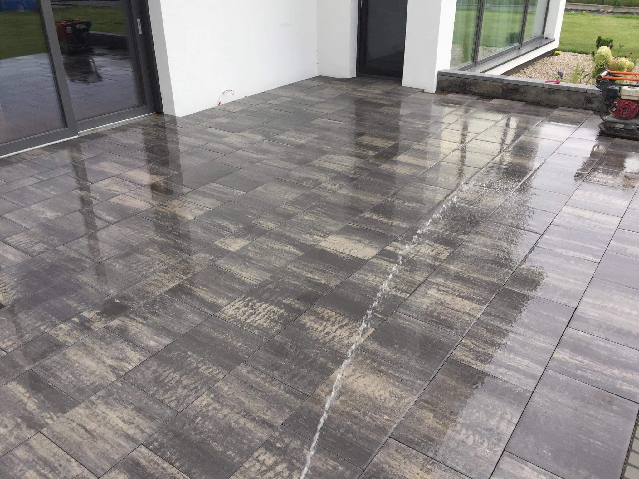 Świeżo ułożona, wilgotna kostka brukowa w odcieniach szarości i beżu przed nowoczesnym budynkiem z dużymi oknami. W tle widoczny trawnik i fragment ogrodu. Woda spływa po powierzchni kostki...