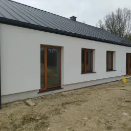 Jasna elewacja domu jednorodzinnego z ciemnym dachem z blachy na tle pochmurnego nieba i drzew bez liści, widoczne brązowe ramy okienne i teren wokół budynku w trakcie prac wykończeniowych.