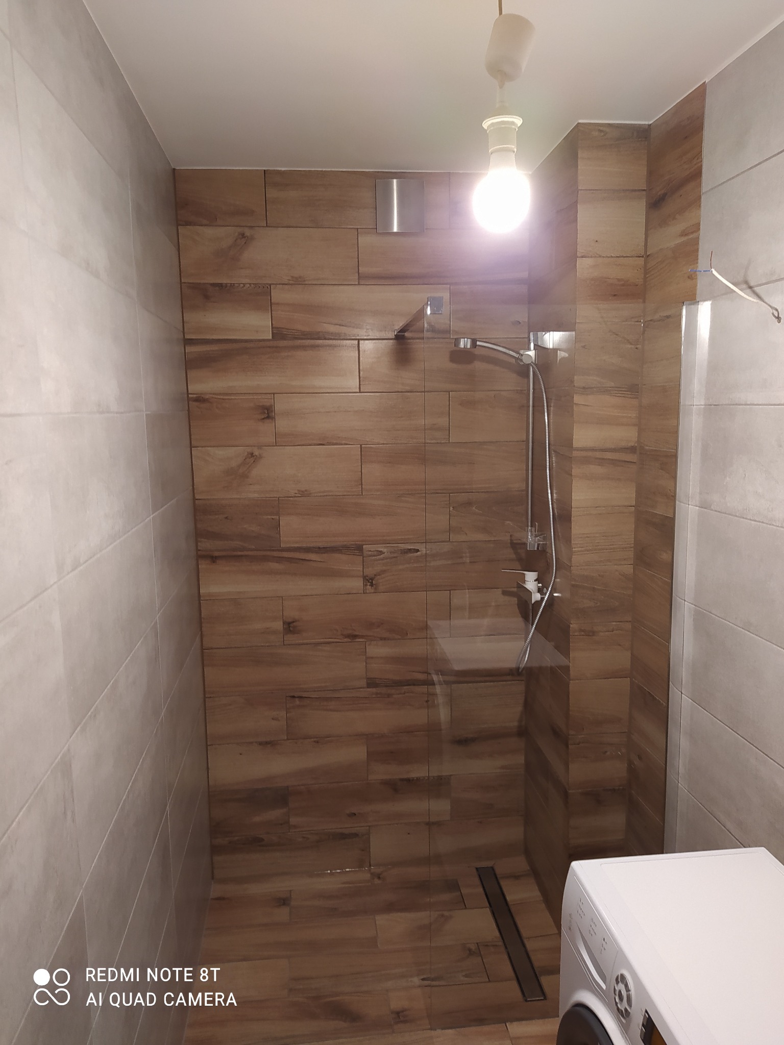 Nowoczesna kabina prysznicowa z odpływem liniowym, wykończona płytkami imitującymi drewno, obok biała pralka.