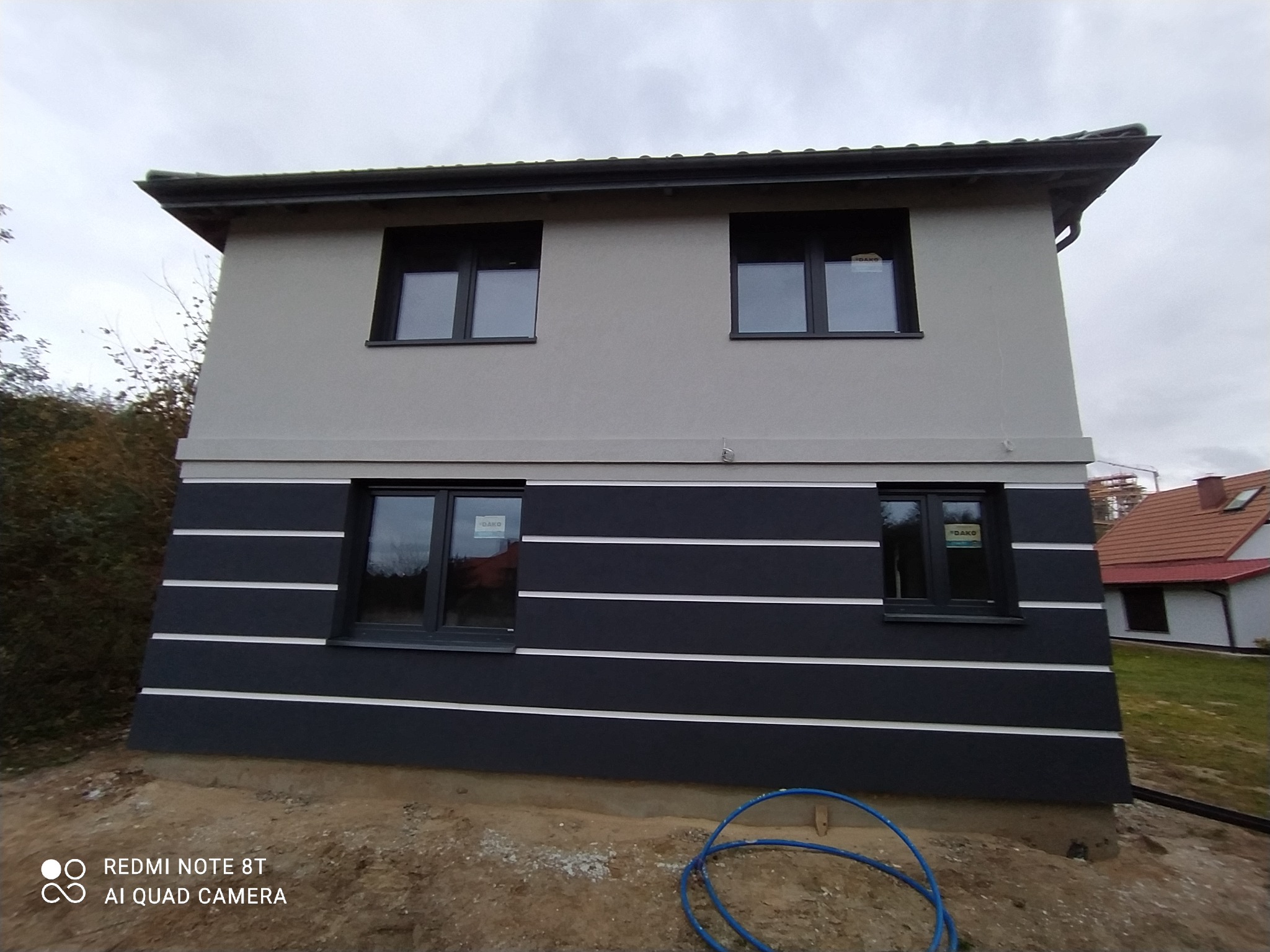 Dwukondygnacyjny dom z elewacją w kolorze szarości na górze i ciemnogranatowym z białymi pasami na dole, widok z zewnątrz, niebieski wąż leżący u podstawy ściany.