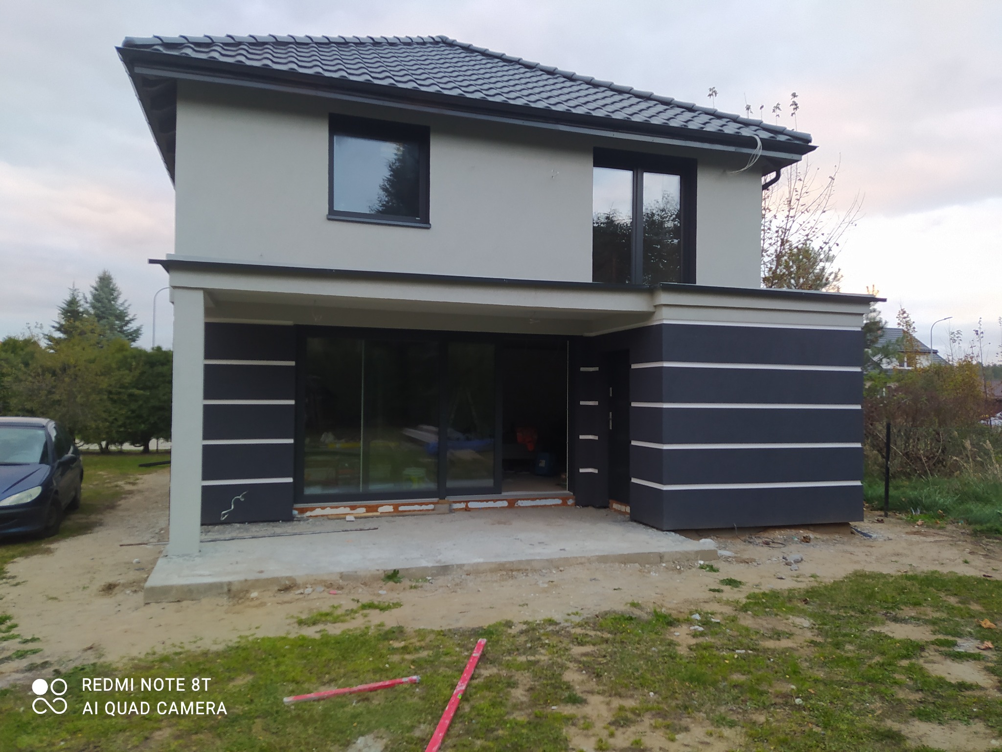 Dwukondygnacyjny dom z szarą elewacją i grafitowymi akcentami, widoczne ślady prac wykończeniowych na zewnątrz, w tle zieleń i fragment zaparkowanego samochodu.