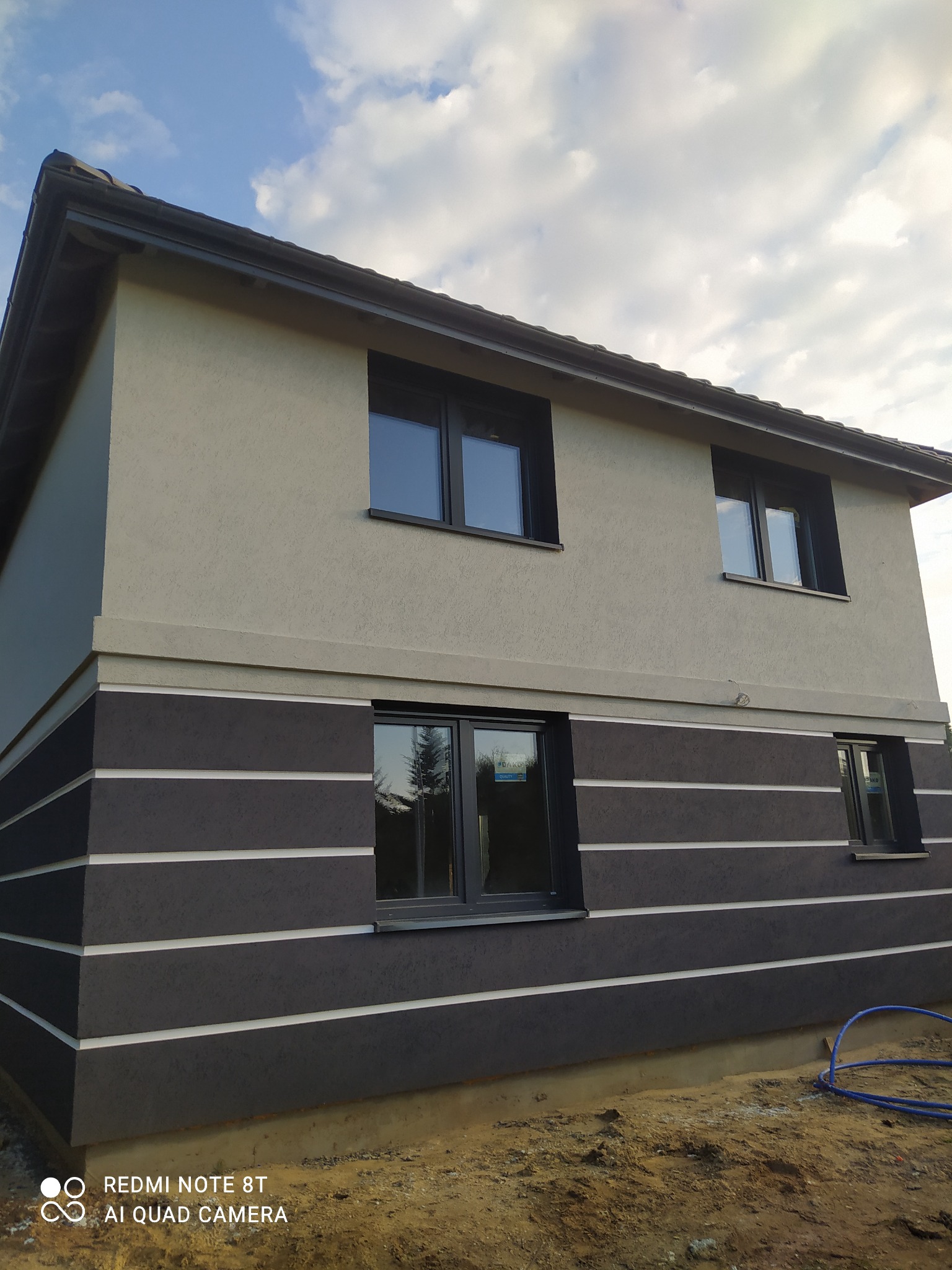 Dwukondygnacyjny dom z elewacją w odcieniach szarości i grafitu, z poziomymi białymi liniami na dolnej części elewacji, widok z dołu na tle pochmurnego nieba.