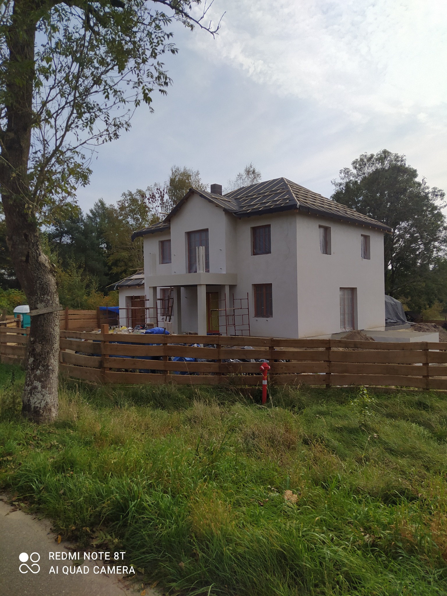 Dom jednorodzinny w stanie surowym zamkniętym w Iławie, z widocznym szkieletem dachu i elewacją przygotowaną do malowania, ogrodzony drewnianym płotem.