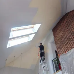 Mężczyzna maluje ścianę w pomieszczeniu ze skosami i oknami dachowymi, stojąc na aluminiowej drabinie.