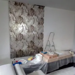 Montaż tapety z motywem liści na ścianie, widoczna drabina i narzędzia malarskie na stole przykrytym folią.