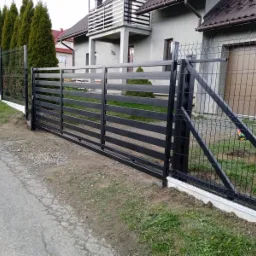 Czarna, pozioma brama wjazdowa z metalowych profili oraz przyległe ogrodzenie z siatki na tle szarego domu z balkonem i tujami.