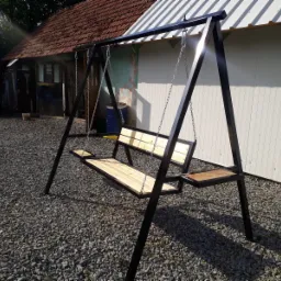 Metalowa huśtawka ogrodowa z drewnianym siedziskiem i bocznym stolikiem, wykonana w Męcinie, na tle budynku z czerwoną dach&oacute;wką i białej ściany.