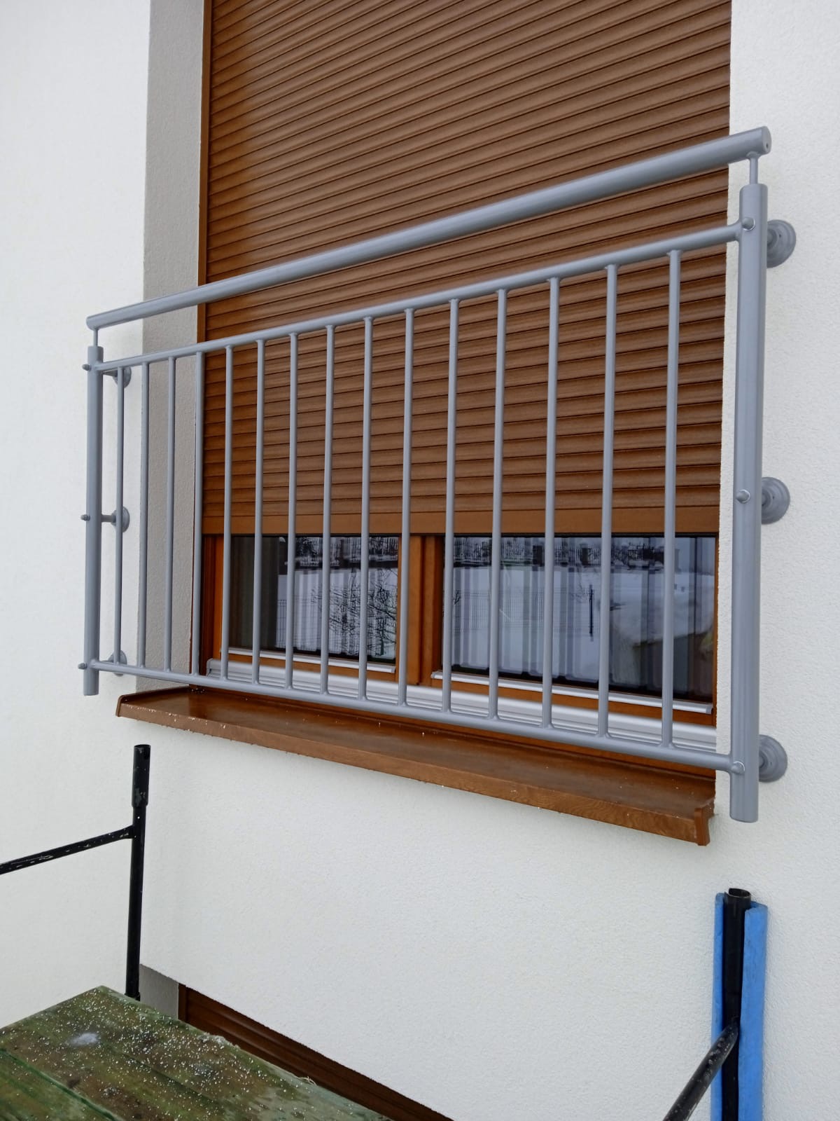 Szara, metalowa balustrada okienna z pionowymi szczebelkami, zamontowana na białej elewacji budynku, widok z zewnątrz, z brązowymi roletami w tle.